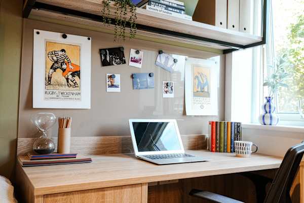 A wide wooden study desk with a laptop, books, pencils, and a mug, set against a pinboard displaying posters and photographs, with a large window to the right letting in natural light.