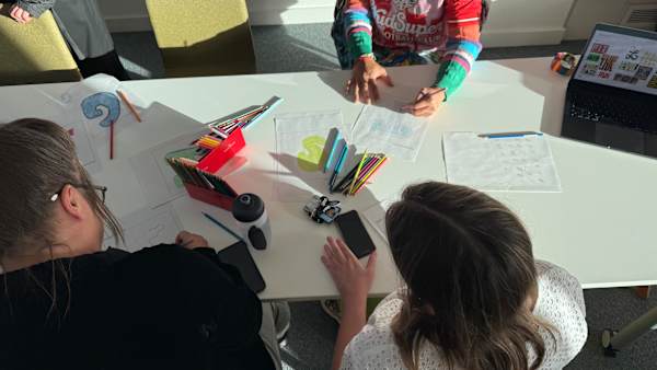 Overhead view of participants sketching colourful letterforms around a table with pencils, papers, and workshop materials.