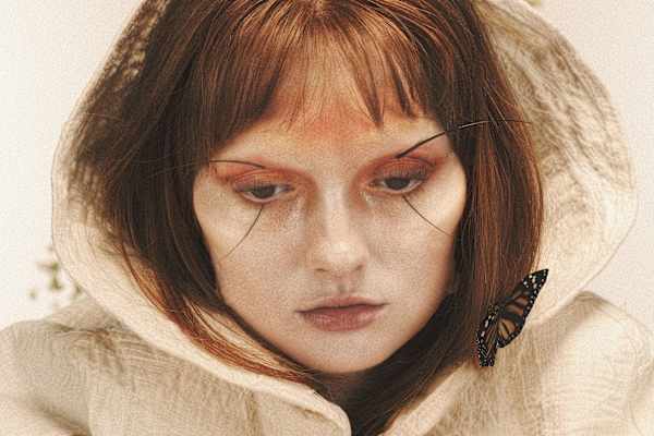 Close-up portrait of a person in a sheer white hood with dramatic graphic eye makeup and a butterfly resting on the shoulder.