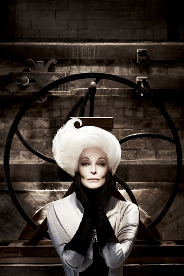 photo of Carmen Dell'Orefice posed head in gloved hands, hair swept upwards, in front of a black wheel sculpture against a brown textured wall designed like an antique box