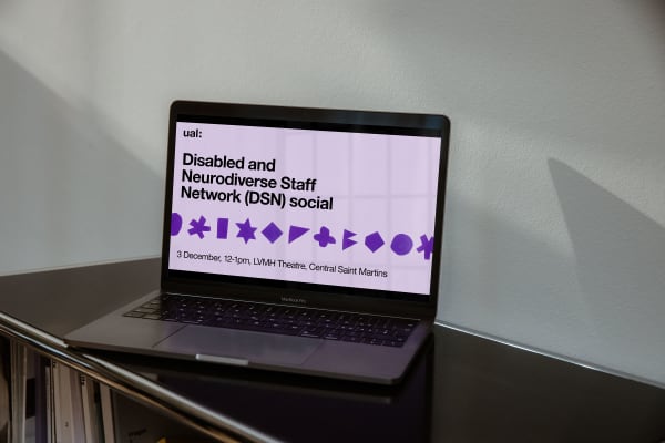 A digital event announcement for the Disabled and Neurodiverse Staff Network social at Central Saint Martins on 3 December at 12pm to 1pm at LVMH Theatre, Central Saint Martins with purple diverse symbols. The announcement is displayed full-screen on a silver laptop. The laptop is sat on top of a short bookshelf and a white painted wall is behind it. 