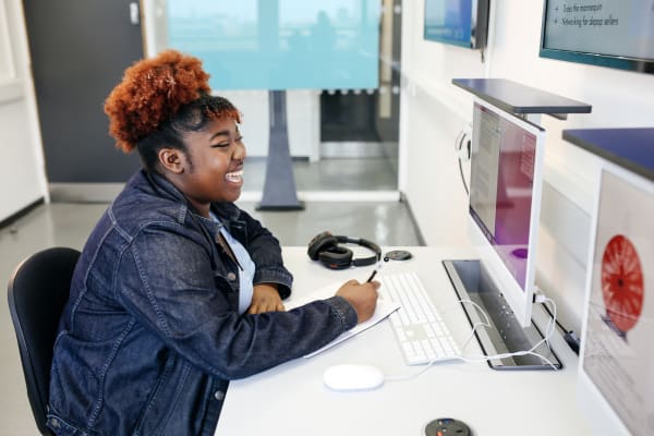 Mo is sitting at a desk facing a computer monitor. They are laughing while they ready through what's on their screen.