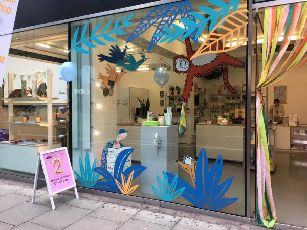 Exterior view of the 'not just a shop' storefront decorated for a celebration. The window features a cheerful jungle-themed display with large, colourful cut-outs of tropical leaves, birds, and a smiling orangutan hanging from vines. A cartoon figure sits among the foliage, reading a book. Blue balloons are attached to the window and an A-board sign on the pavement reads: 