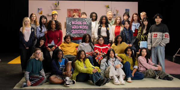 A large, diverse group of approximately 25 young people — students of various ethnicities — are arranged in three rows on a studio stage set. The backdrop is a dusty pink flat decorated with small framed artworks, shelves with ceramics, and dried flower arrangements, suggesting a lived-in domestic space. In the centre of the group, one person holds up a hand-painted chalkboard sign in pink graffiti-style lettering that reads: 