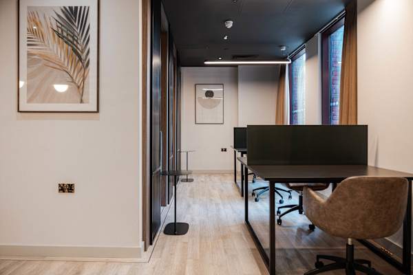 A study area with desks and desk chairs, a lamp and windows. The walls are painted white and there is wood flooring.
