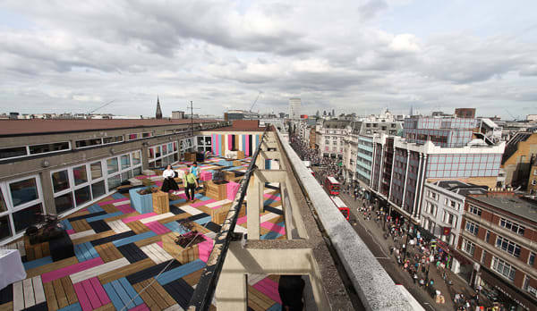 skyline photo of the roof garden- people walking across, blue, pink, white, brown and black patterened floor with similar coloured benches of various sizes. Beneath the roof garden we see the busy london cityscape with pedestrians and buildings.