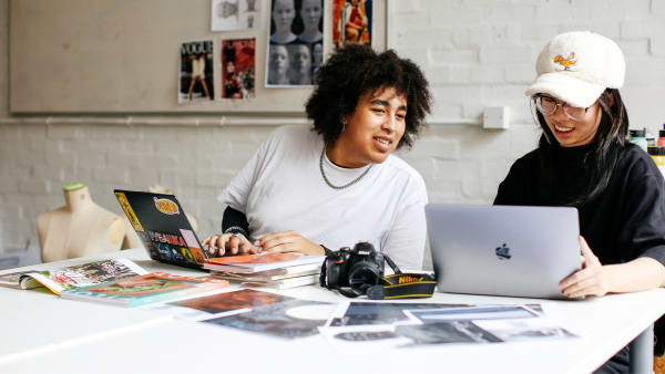 Two students sit at a table scattered with magazines, working on their laptops. They are smiling and collaborating. 