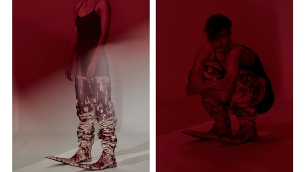 A person in a black tank top and abstract print pants stands and squats in a red-lit space. The mood is artistic and avant-garde.