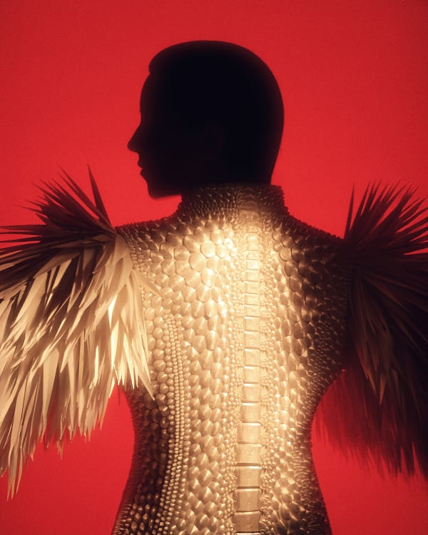 Person wearing a white jacket with sharp spikes looking to the left against a red background.