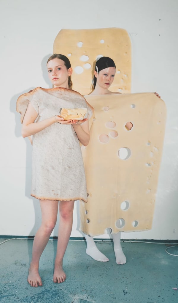 Two models stand together, one wearing and the other holding out in front of them, garments resembling a slice of ham and a slice of swiss cheese. Another slice of cheese is suspended behind them both.