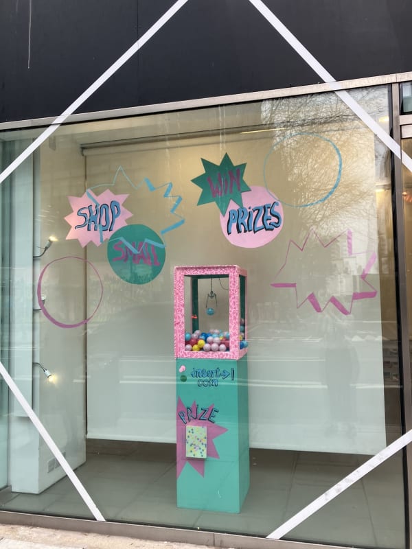 A playful window display at 'not just a shop' featuring a colourful claw machine installation. The machine is decorated in turquoise, pink, and patterned textures, filled with pastel plastic balls and a hanging claw. Hand-painted signs on the glass in comic-style bursts read: “SHOP SMALL” and “WIN PRIZES” in bold pink, green, and blue lettering. Additional circular and starburst shapes are scattered across the glass for a dynamic effect. The display promotes an interactive and fun shopping experience, visible from the street through the large front window.