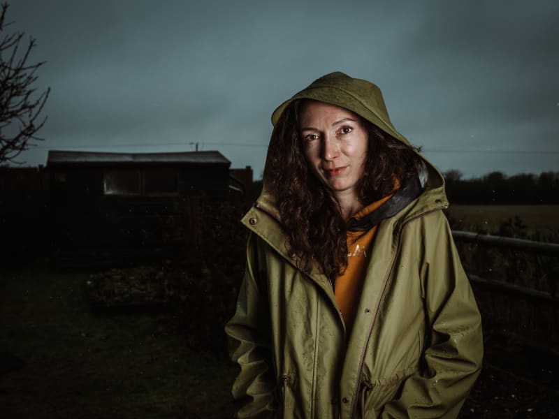 A women called Anzhela is stood outside in the rain, looking straight at the camera with her hands in her raincoat. 