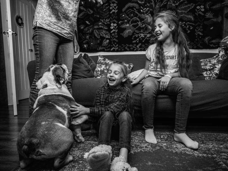 Two young girls are sat on the floor and sofa playing with their dog. 