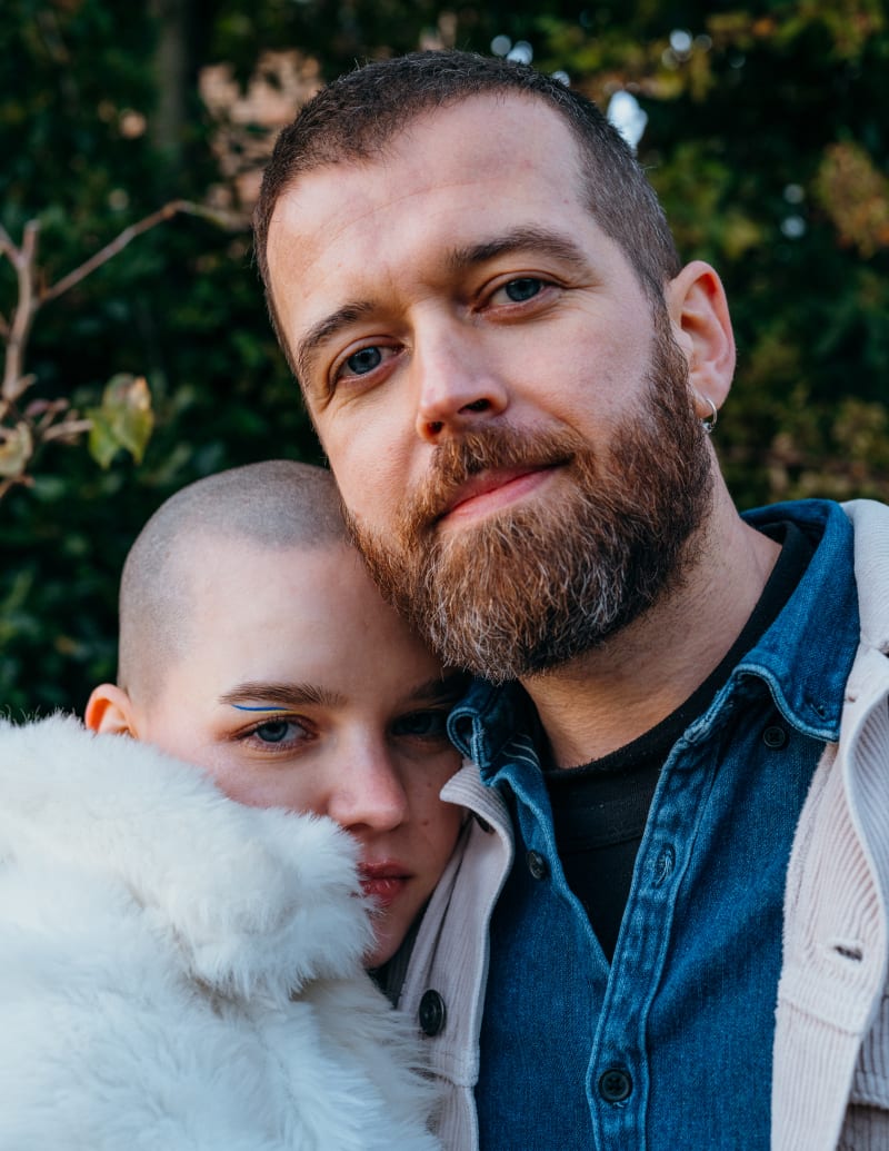 A young couple look directly at the camera. The women's head in the shoulder of the mans. 