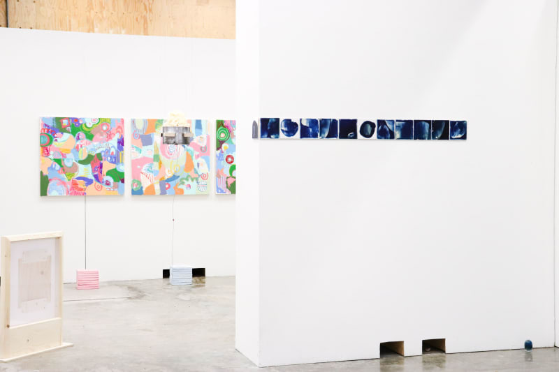 Twelve small ceramic tiles are arranged horizontally on a white wall. The surface of the tiles is coated in a blue bioplastic and images of the body.