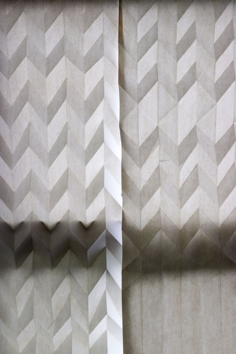 2 sheets of geometrically folded paper hung against window. Light shines through the paper, emphasises the folds.