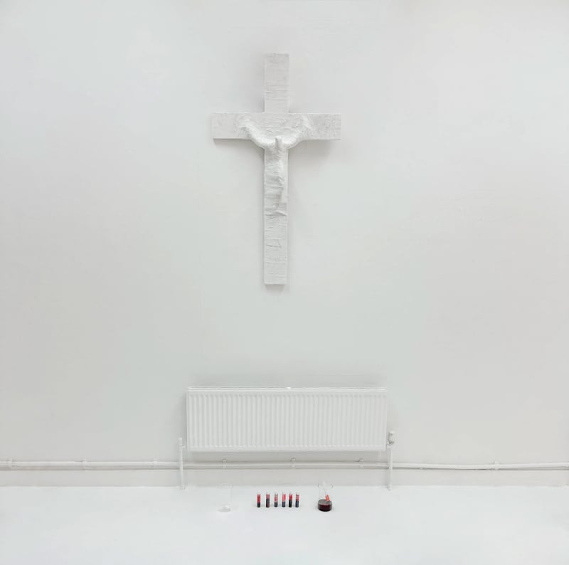 Image of a crucifix covered by plaster on a white wall that is gradually covered in red paint by the public.