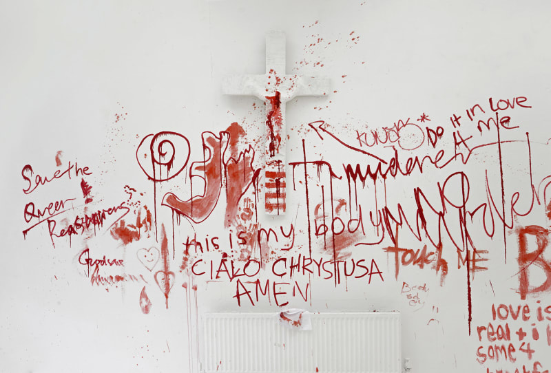 Image of a crucifix covered by plaster on a white wall that is gradually covered in red paint by the public.