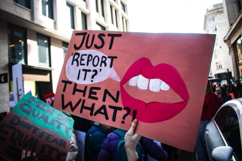 A handmade cardboard sign reading: Just 'Report It?' Then What?! with a speech bubble and a mouth painted onto the sign. 