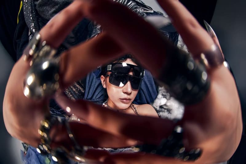 A woman wearing oversized sunglasses and heavy jewellery frames their face with outstretched hands.