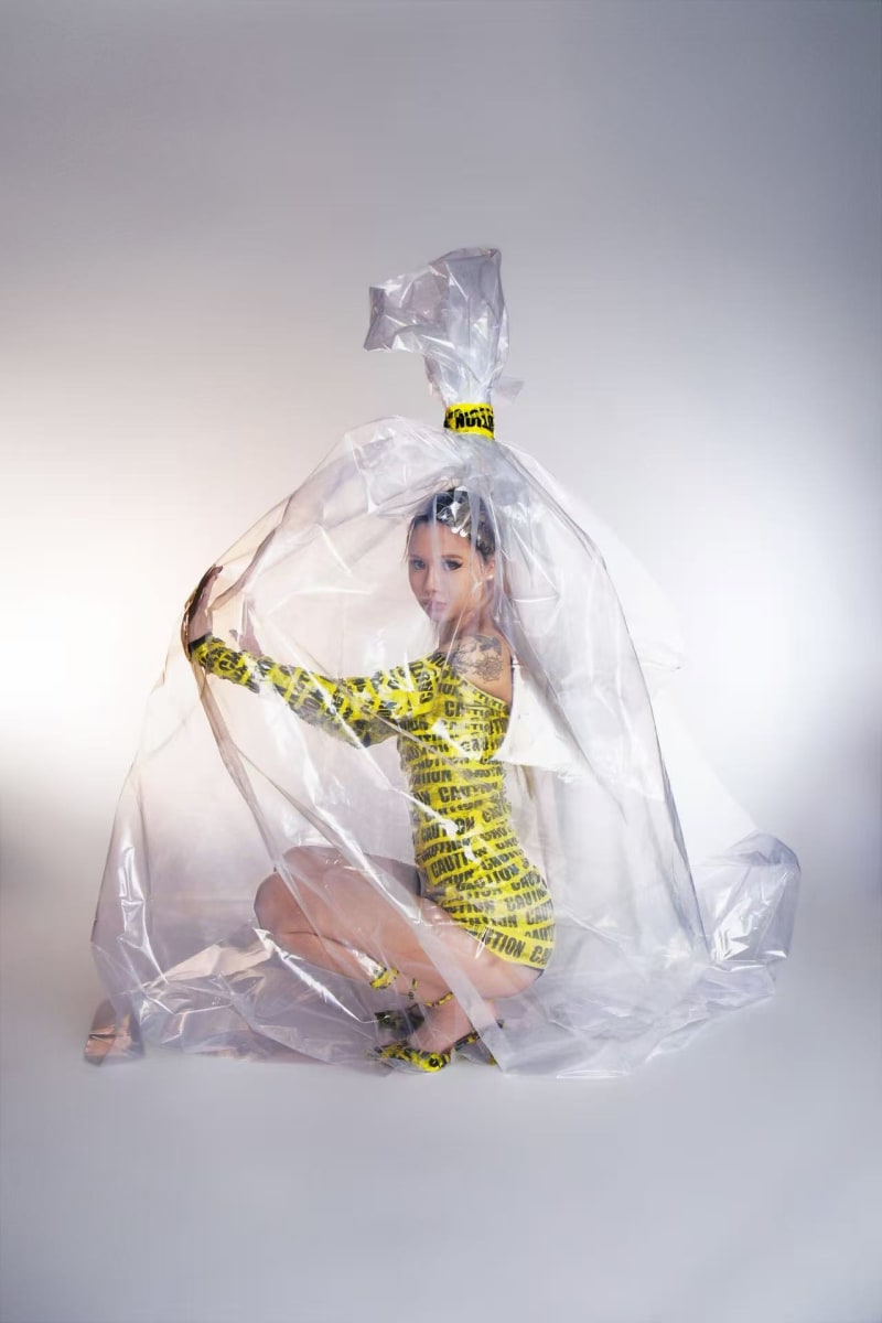 A woman dressed in caution tape is crouched inside a large, tied plastic bag.