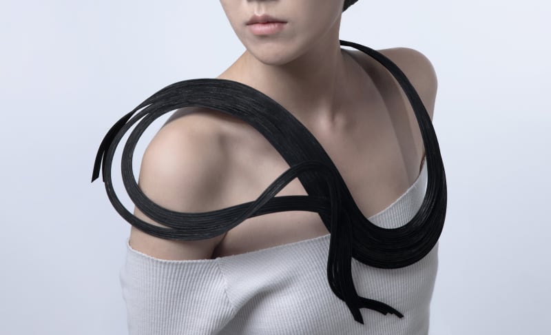 A model wears a black rattan chest ornament. The piece has a fluid, curved design that wraps smoothly around the shoulder and neck.