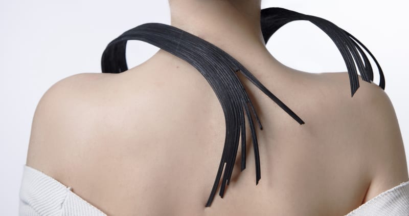 A model wears a black rattan chest ornament. The piece has a fluid, curved design that wraps smoothly around the shoulder and neck.