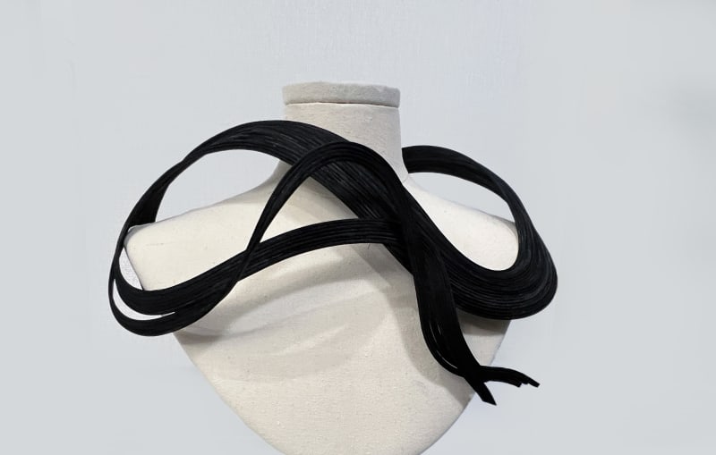 A model wears a black rattan chest ornament. The piece has a fluid, curved design that wraps smoothly around the shoulder and neck.