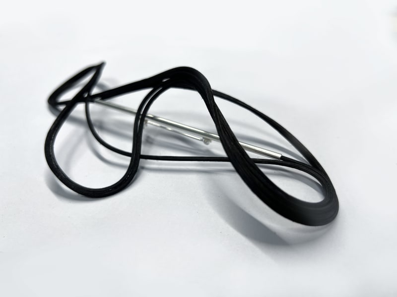 The brooch features rattan and silver tubes. The rattan pierces through the silver tubes and wraps around them, forming a smooth, flowing design aroun