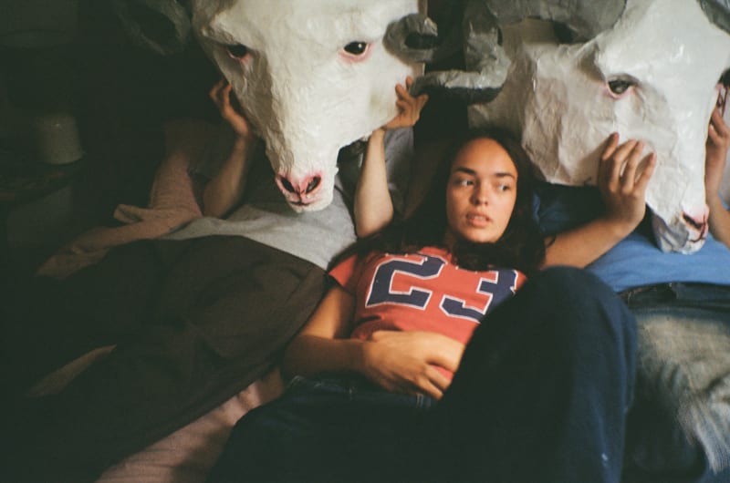 Two boys with large ram masks on a bed with a girl in a series of film photography.