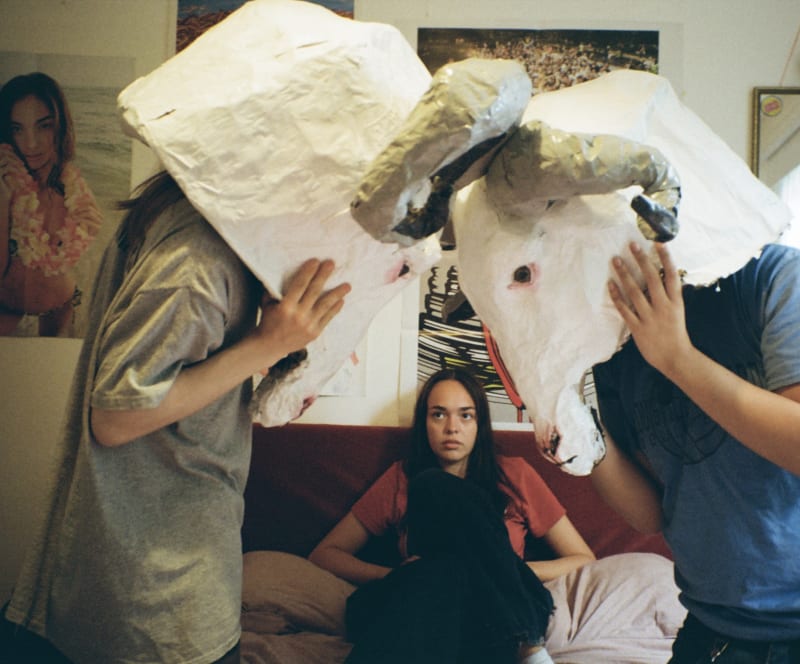 Two boys with large ram masks on a bed with a girl in a series of film photography.