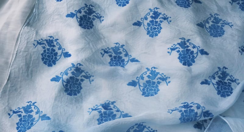 A pale blue silk scarf with soft, flowing gradients, dyed using phycocyanin pigment. It features repeating lotus-inspired patterns, simplified from tr