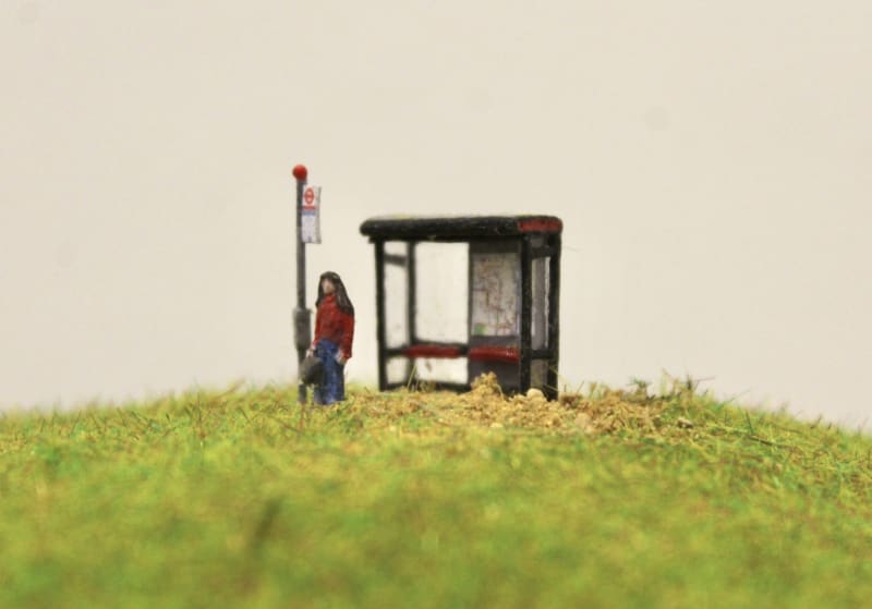 A miniature model of a person in a red jumper, blue jeans and black boots waiting at a miniature bus stop. 