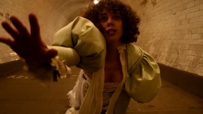 A woman in a Victorian inspired costume reaches out from the floor of a dimly lit tunnel