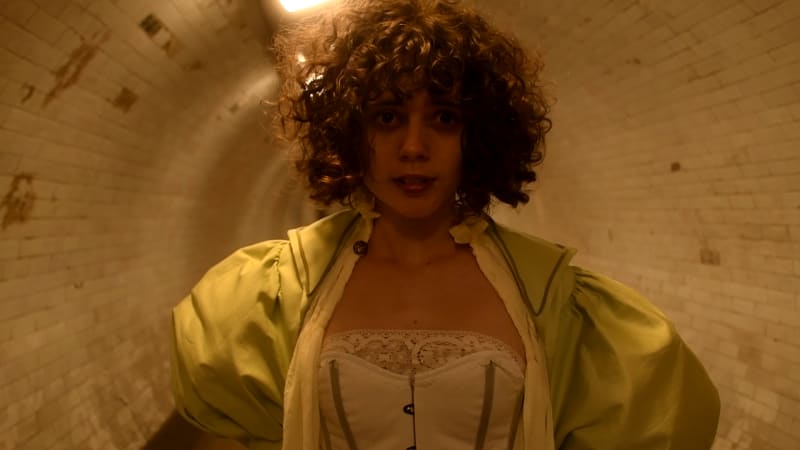 A close up of woman in a Victorian inspired costume standing, frightened, in a dimly lit tunnel.