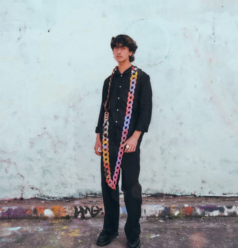 A man wearing a big necklace with colourful paint on stand infront of a wall.