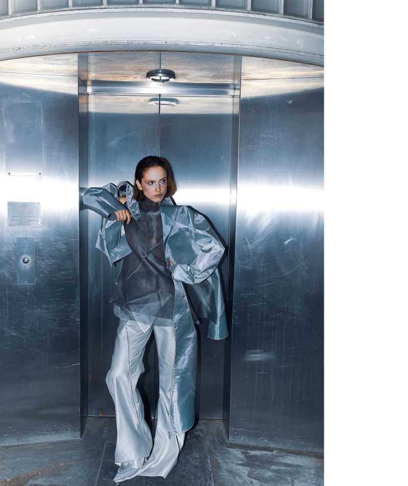 Female model in a shiny black sculptural coat stands on an outdoor concrete stairway, her figure split between deep shadow and bright sunlight.