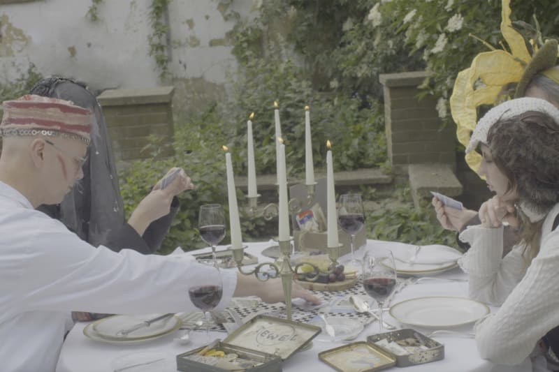 Characters playing a card game around a table in a garden, wearing knitted headpieces