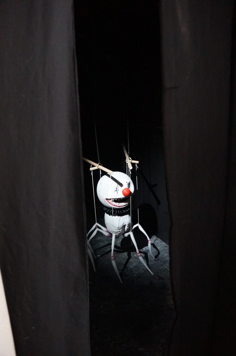 3D printed, six legged creature puppet with white clown makeup and a red nose seen through the gap of black curtains hanging in a doorframe.