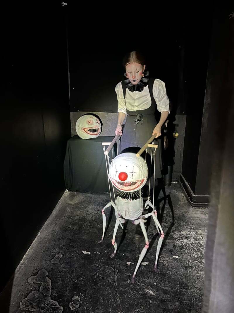 Person dressed in a black and white clown outfit, with clown makeup, holding a six legged creature puppet in a black room.