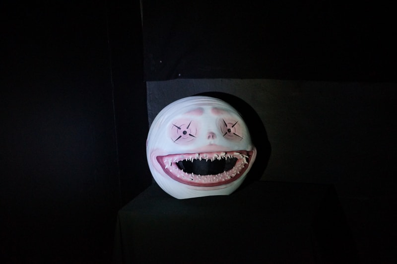 A big mask head with a huge grin, lots of sharp teeth and beady eyes on a black plinth.