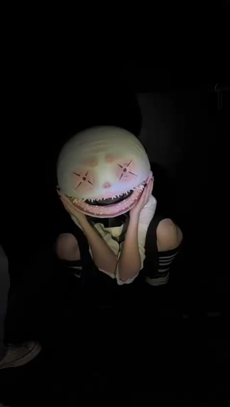 Person dressed as a clown, crouched on the floor in a black room wearing a big smiling mask head