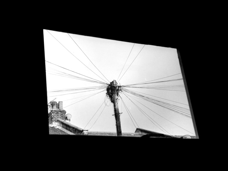 Black and white image taken out a window. Framed by the window is a powerline in-front and the power lines are spread across the entire frame.