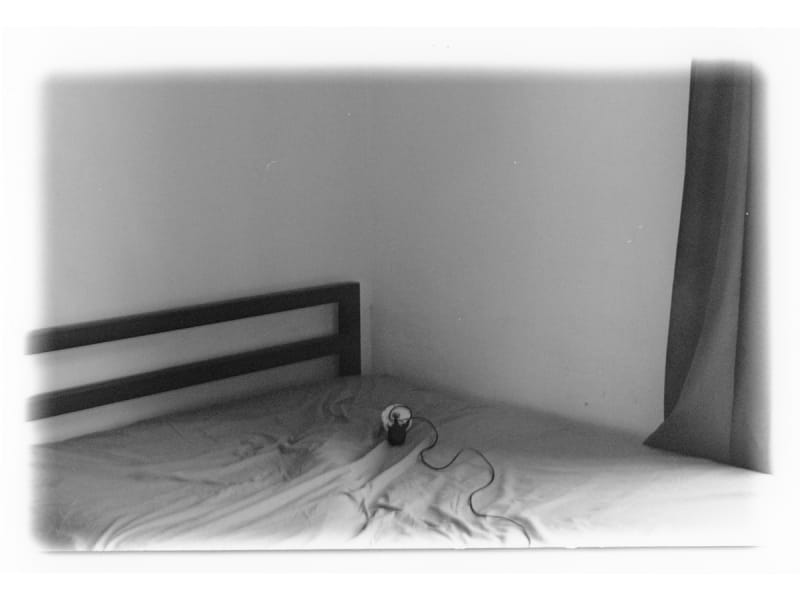 Black and white of a bed in the corner of the room with cable release where someone should be sat.