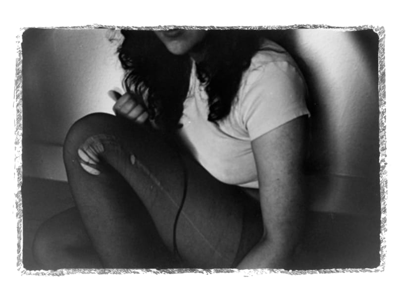 Black and white image of young woman curled up on the floor, focus on the ripped tights she is wearing. The image is cropped to cut her face out.