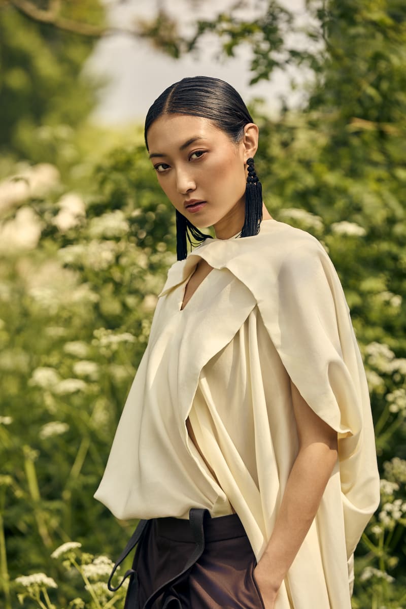 Medium shot of asian female model wearing white satin layered top and brown trousers with outdoor greenery setting.