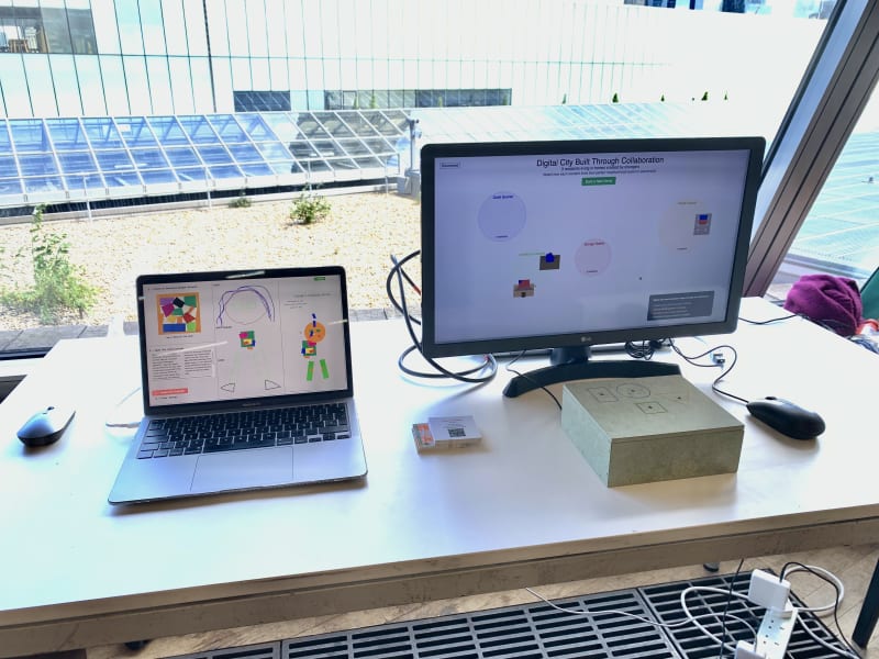 Two monitors on a table by a large window. One laptop shows a colourful AI-generated figure made from selected shapes; the second screen displays the 