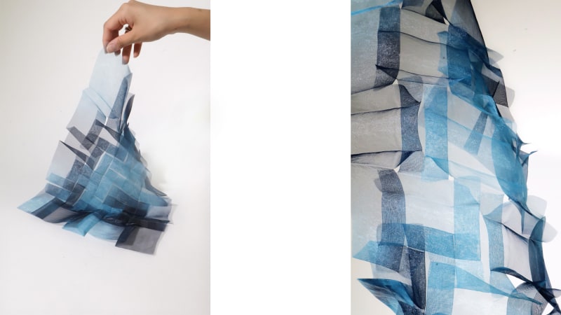 Folded translucent textile in blue and deep navy tones, forming a flowing architectural shape as it's gently lifted by hand.