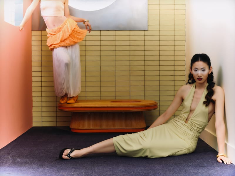 Two women pose in a staged tiled interior, alternating between standing and reclining, highlighting their outfits through minimal geometric settings.