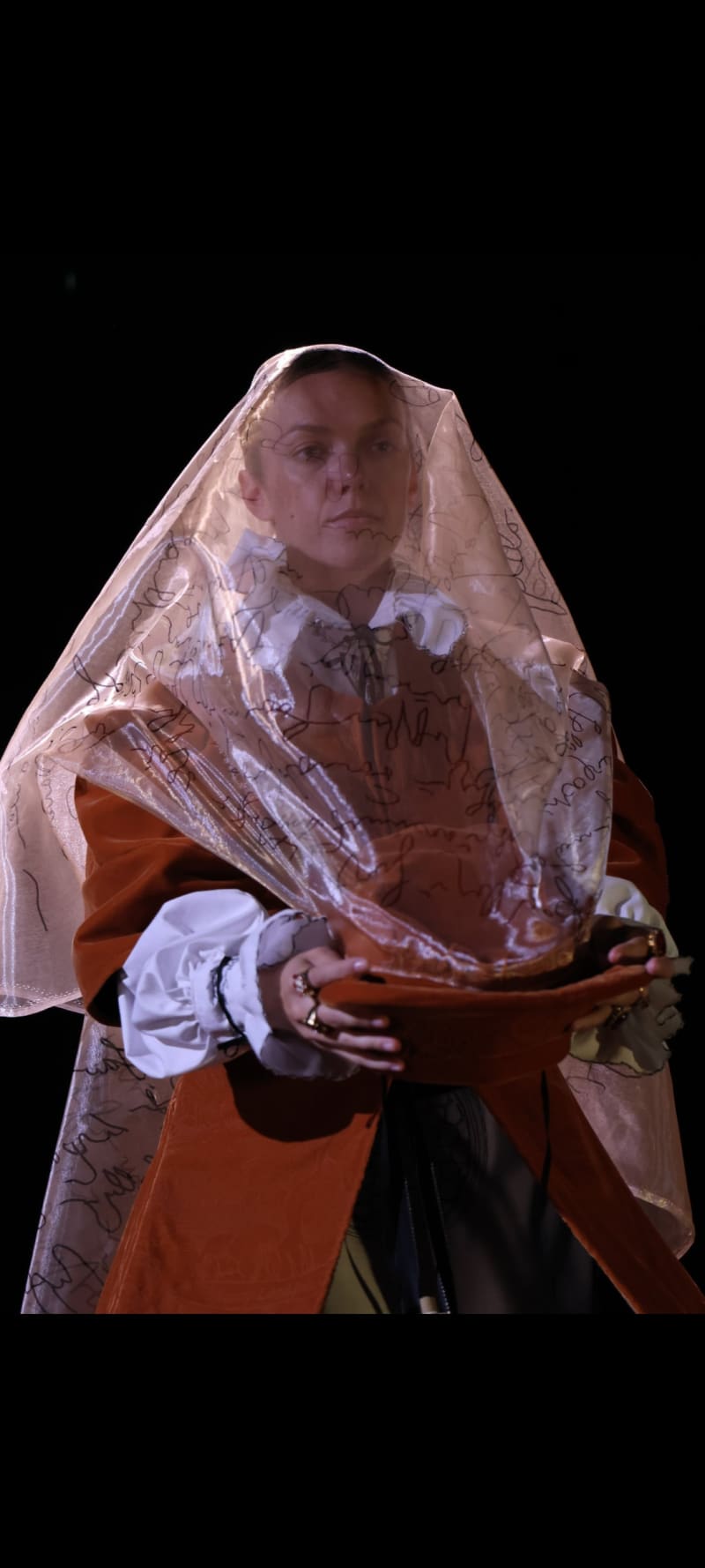 Image of a woman pulling a veil over her face. The veil has 16th-sentury script on it.